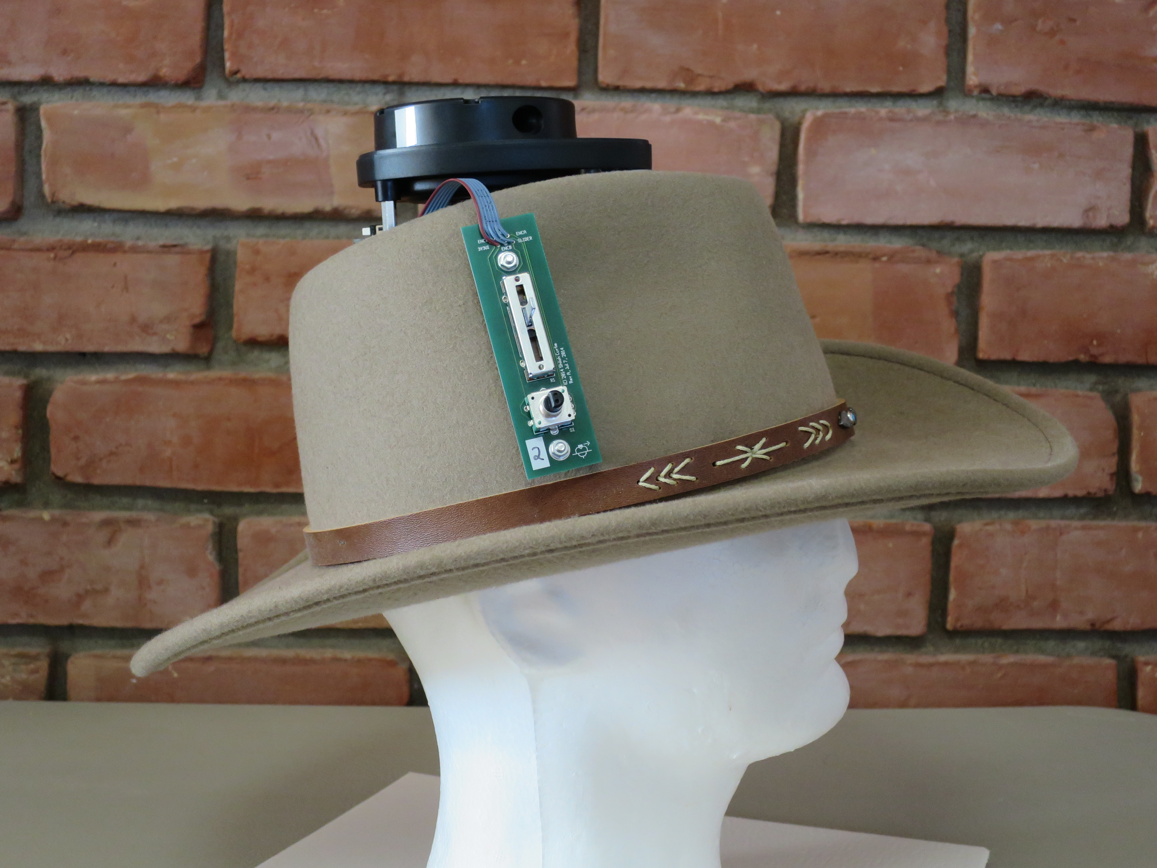 Picture of the H-NAV.  It’s a light brown cowboy hat with a LiDAR mounted on the top and a PCB with a knob and slider on the side.  It is sitting on a white foam display head.